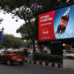 A realistic urban DOOH billboard displaying a Coca-Cola advertisement on a busy city roadside with moving traffic, trees, and modern outdoor advertising infrastructure.