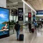 Travelers walking through a modern airport terminal surrounded by premium digital advertising displays and large-format airport OOH campaigns promoting travel, luxury, and lifestyle brands.