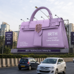 A realistic oversized lavender luxury handbag installation by IRTH displayed along Mumbai’s Western Express Highway, surrounded by moving traffic and urban skyscrapers, showcasing an eye-catching premium outdoor advertising campaign.